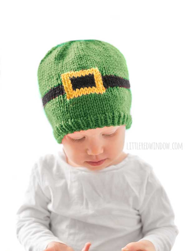 baby in white shirt wearing a green knit hat with a black and gold leprechaun belt around the middle in front of a white background looking down at their lap