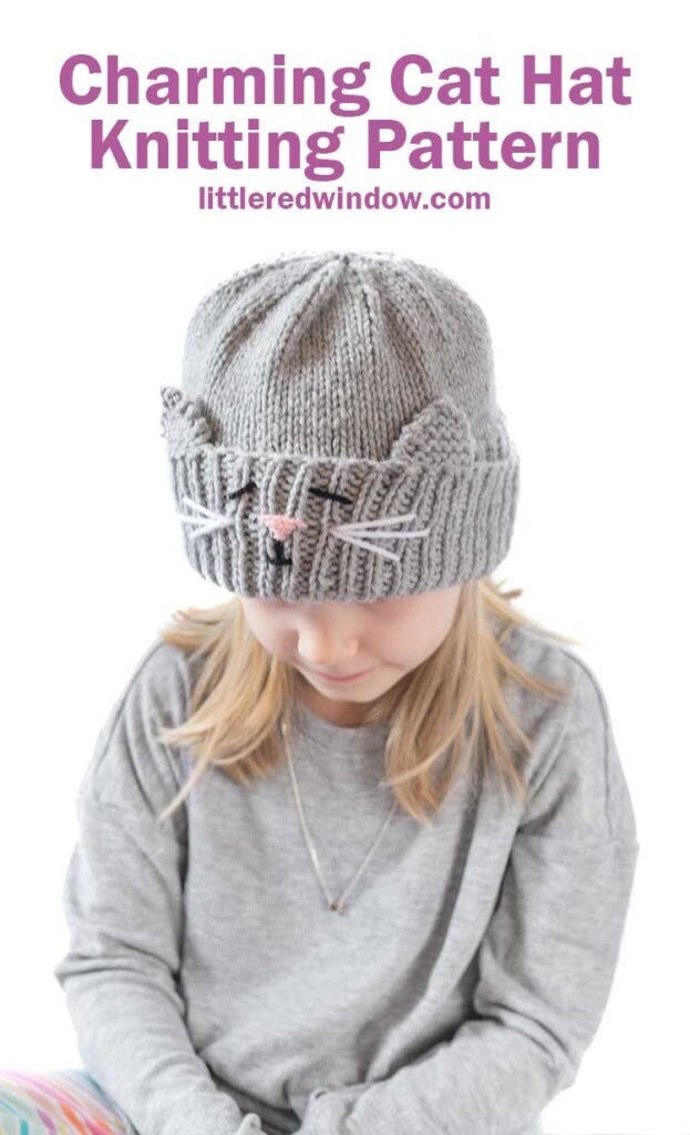 blond girl in gray shirt looking down at her lap and wearing a gray knit hat with a cat face and ears embroidered into the thick ribbed folded brim
