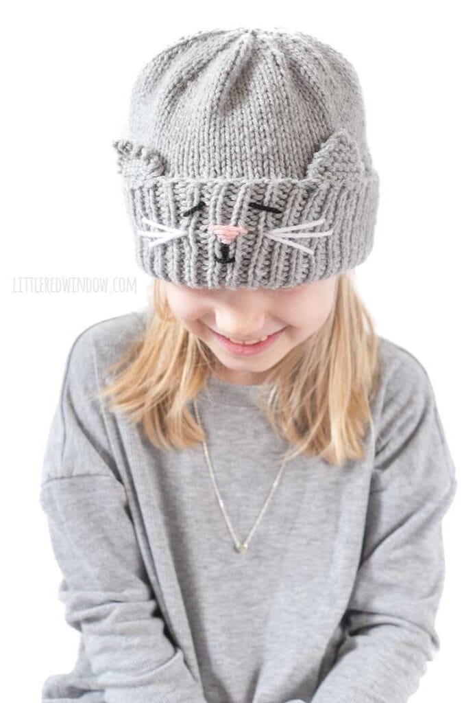 Smiling blond girl in gray shirt looking down at her lap and wearing a gray knit hat with a cat face and ears embroidered into the thick ribbed folded brim