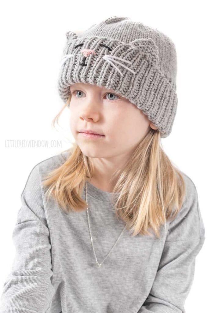 blond girl in gray shirt looking off to the left at her lap and wearing a gray knit hat with a cat face and ears embroidered into the thick ribbed folded brim