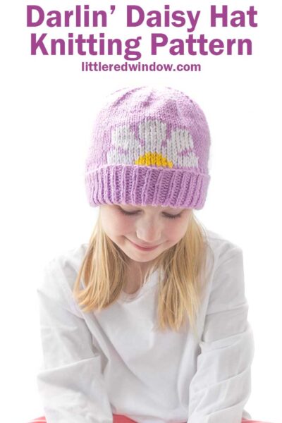 Smiling blonde girl looking down while wearing a white shirt and a lavender knit hat with thick folded brim that has a white and yellow daisy embroidered on the front peeking halfway over the brim