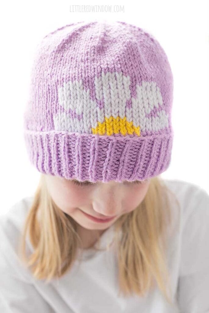 Closeup of Smiling blonde girl looking down while wearing a white shirt and a lavender knit hat with thick folded brim that has a white and yellow daisy embroidered on the front peeking halfway over the brim