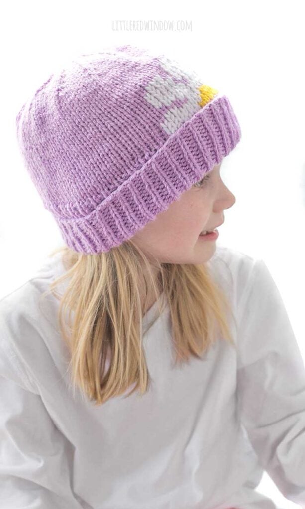 Smiling blonde girl looking off to the right while wearing a white shirt and a lavender knit hat with thick folded brim that has a white and yellow daisy embroidered on the front peeking halfway over the brim