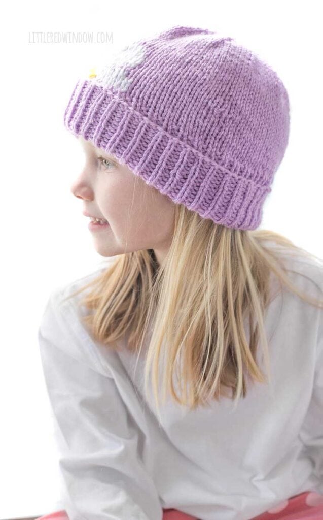 Smiling blonde girl looking off to the left while wearing a white shirt and a lavender knit hat with thick folded brim that has a white and yellow daisy embroidered on the front peeking halfway over the brim