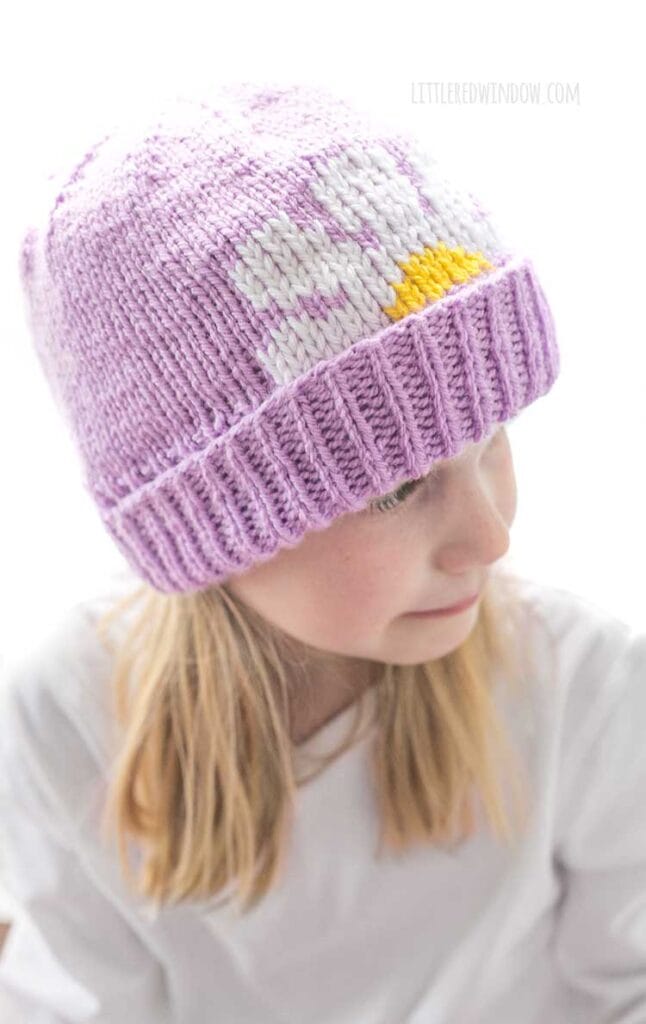 Slightly above view of blonde girl looking off to the right while wearing a white shirt and a lavender knit hat with thick folded brim that has a white and yellow daisy embroidered on the front peeking halfway over the brim
