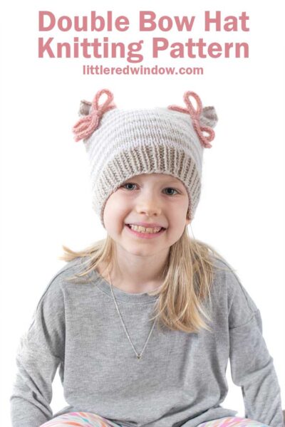 smiling girl in gray shirt looking at the camera and wearing a tan and white thin stripe knit hat that has two pink tied bows at the top that look like ears