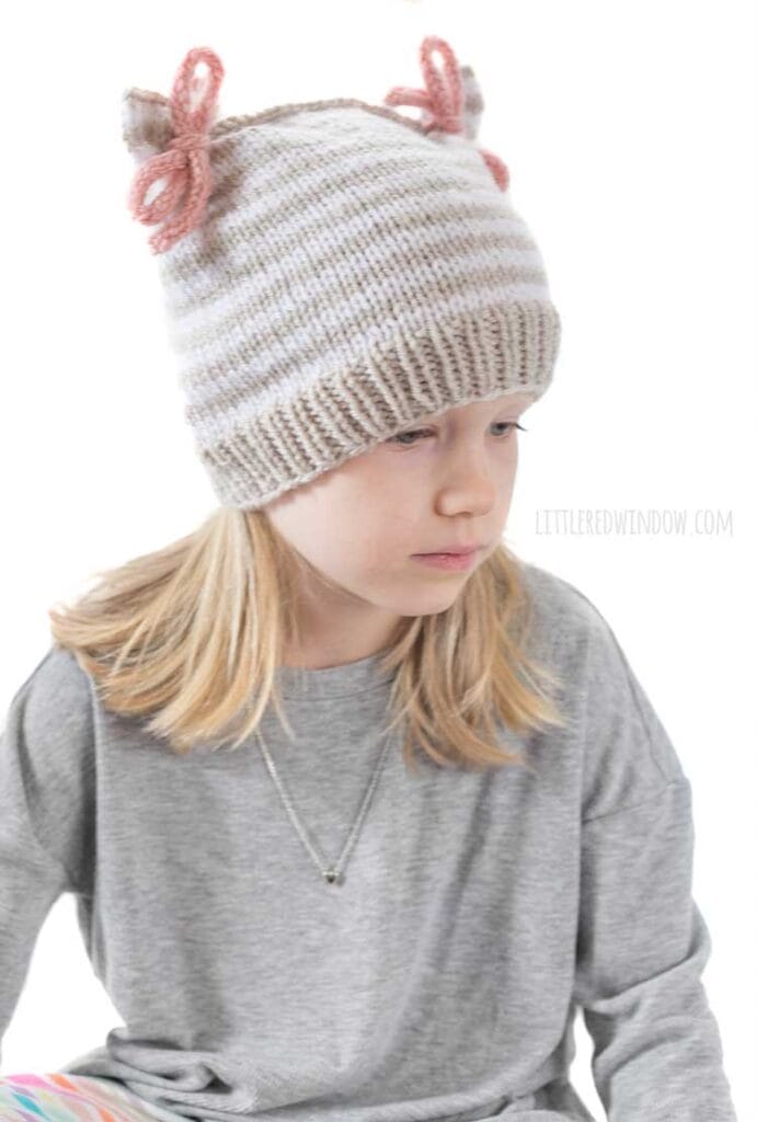 girl in gray shirt looking down to the right and wearing a tan and white thin stripe knit hat that has two pink tied bows at the top that look like ears