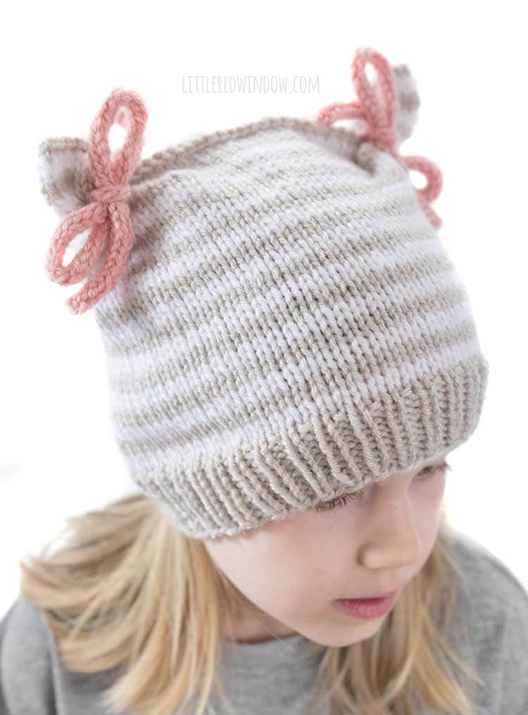 Close up of double bow hat knit in tan and white stripes with two pink bows tied at the top corners