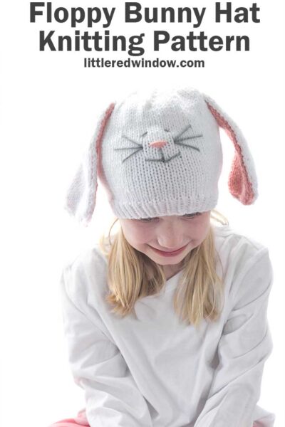 Smiling girl in white shirt wearing a white knit hat with long floppy white bunny ears with pink lining and a cute bunny face on the front looking down at her lap