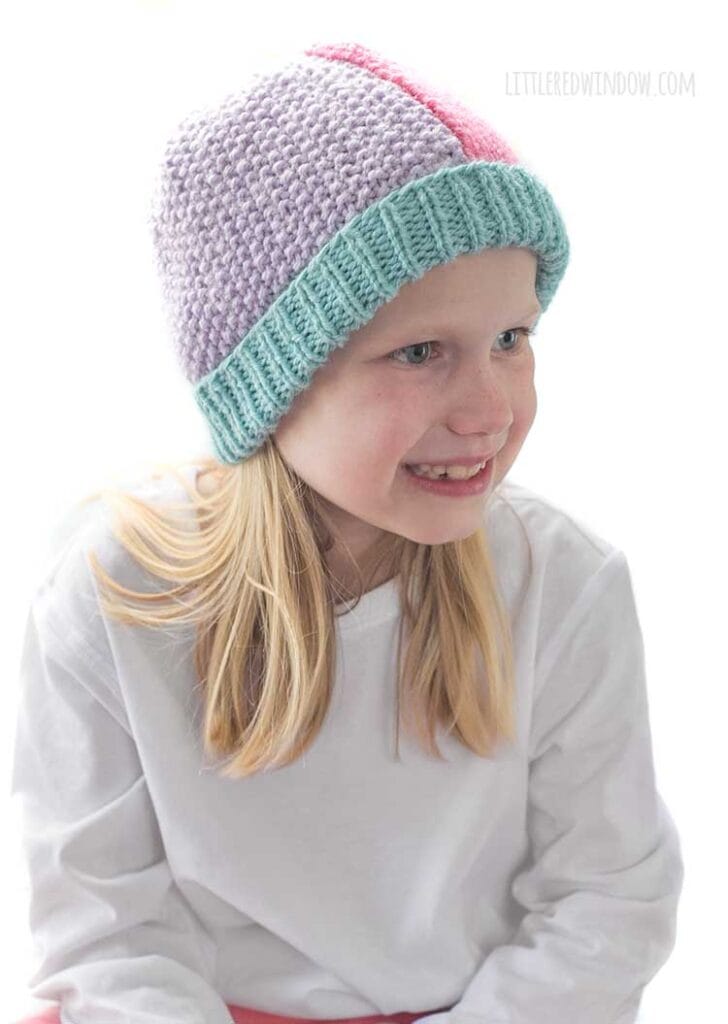 Smiling girl in white shirt looking off to the near right and pink pants wearing a knit hat that has a light teal ribbed brim and is half pink stockinette stitch on the right and half light purple seed stitch on the left
