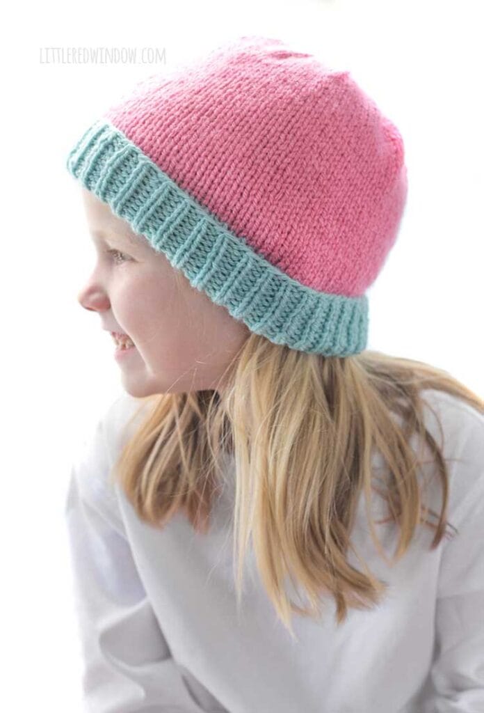 Smiling girl in white shirt looking off to the far left and pink pants wearing a knit hat that has a light teal ribbed brim and is half pink stockinette stitch on the right