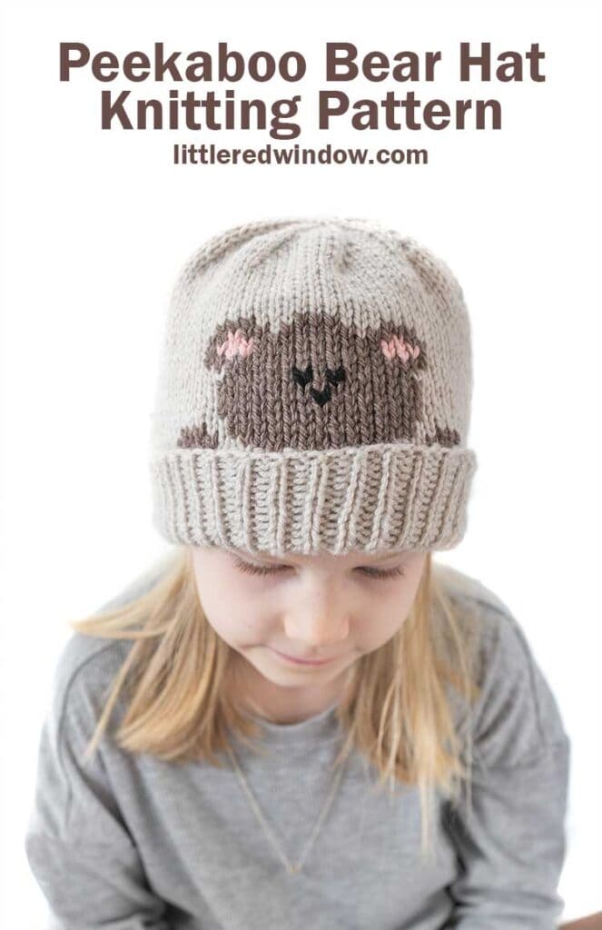 blonde girl in gray shirt looking down wearing a tan knit hat with a folded brim that has a brown teddy bear head peeking over the brim
