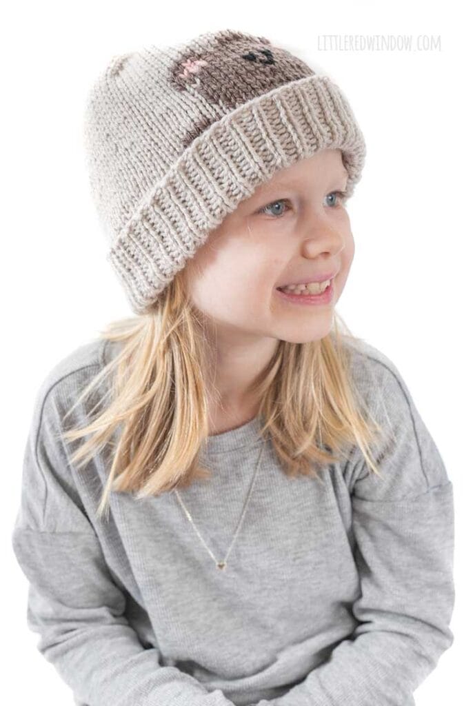 smiling blonde girl in gray shirt looking to the right wearing a tan knit hat with a folded brim that has a brown teddy bear head peeking over the brim