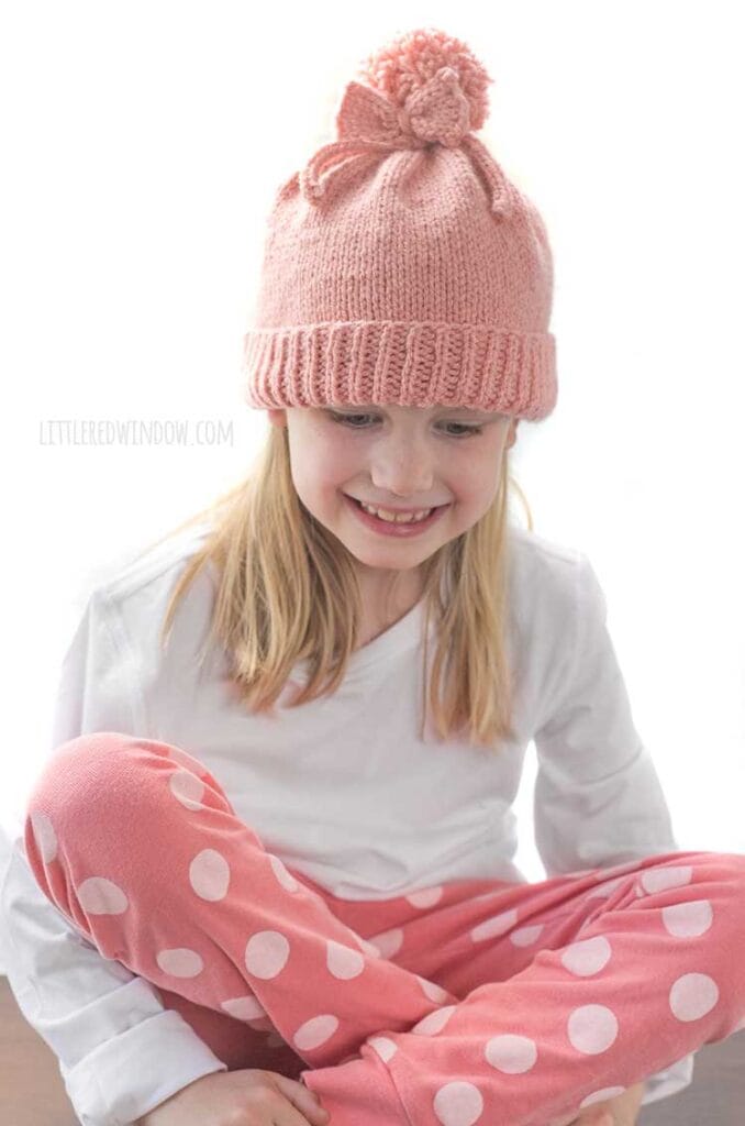 girl in white shirt and pink pants sitting cross legged smiling and lookind down and wearing a medium pink knit hat with a folded brim a big pom pom on top and a bow tied around the pom