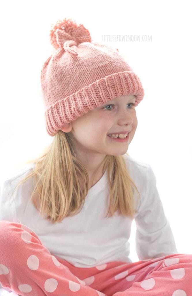girl in white shirt and pink pants sitting cross legged looking off to the near right and wearing a medium pink knit hat with a folded brim a big pom pom on top and a bow tied around the pom