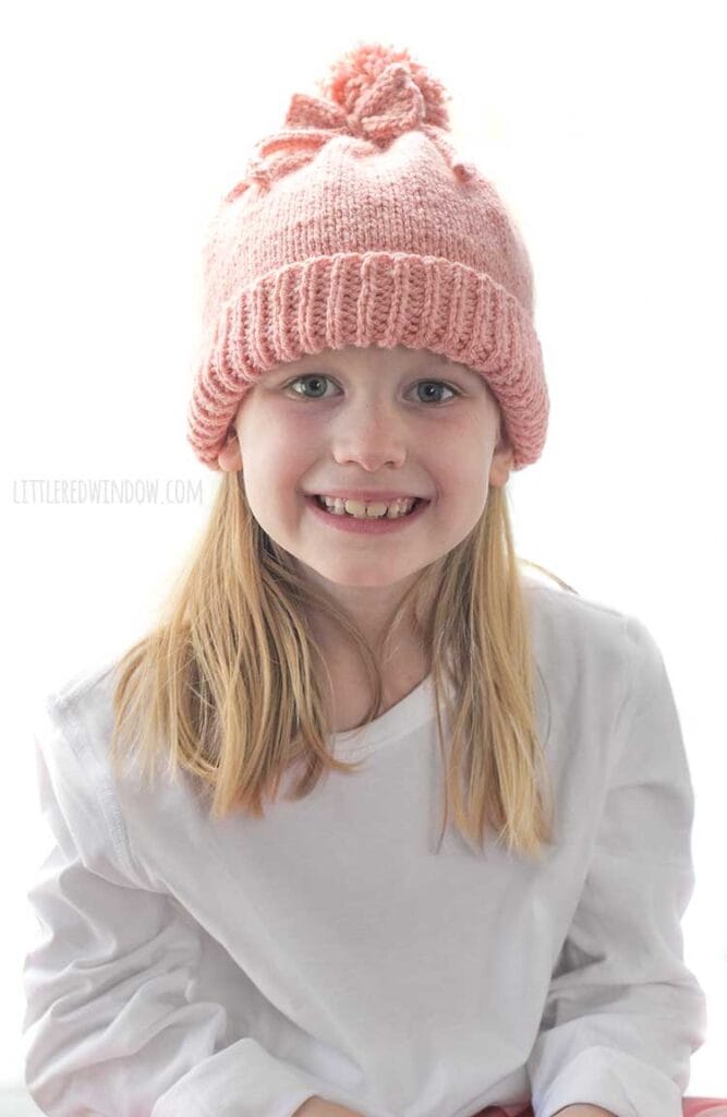 girl in white shirt smiling at the camera and wearing a medium pink knit hat with a folded brim a big pom pom on top and a bow tied around the pom