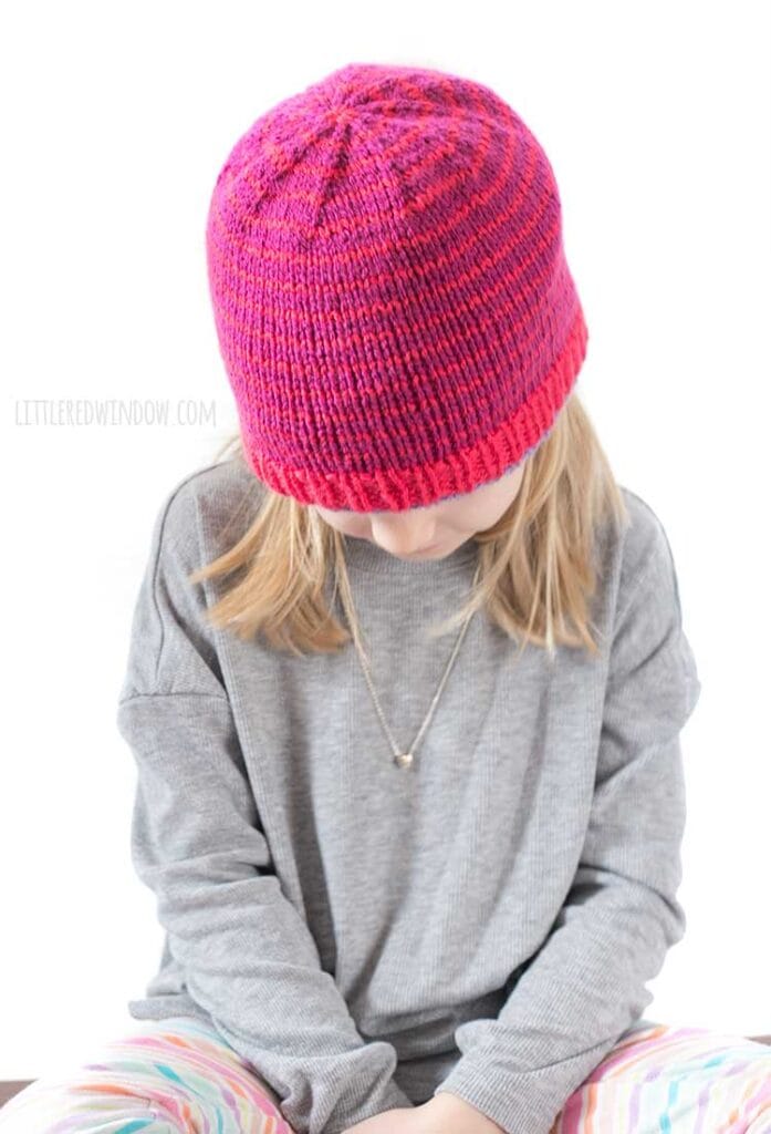 blonde girl in gray shirt looking down at her lap wearing a magenta knit hat with thin purple stripes