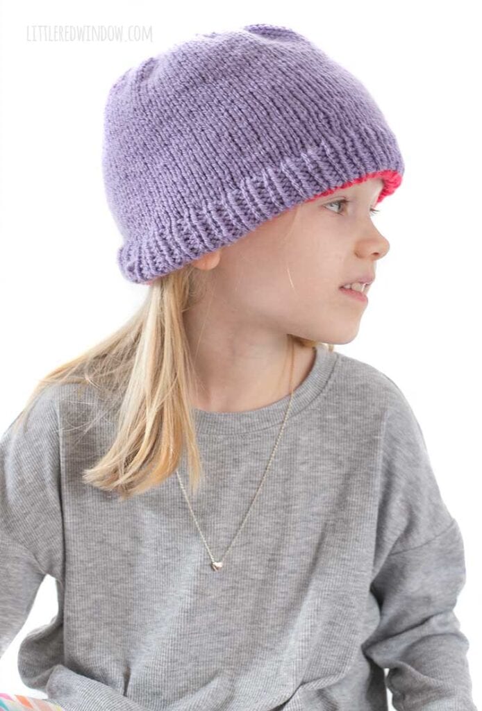 blonde girl in gray shirt looking off to the right wearing a solid light purple knit hat with the magenta reverse color peeking out around the bottom
