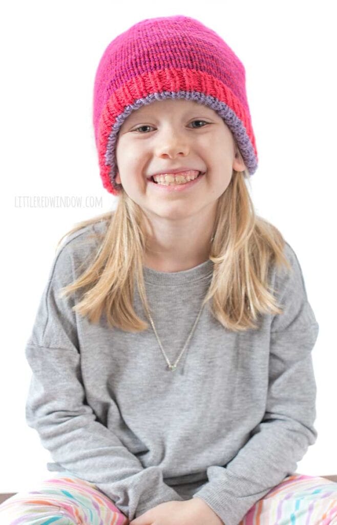 Smiling blonde girl in gray shirt looking right at the camer wearing a magenta knit hat with thin purple stripes with the light purple alternate side of the hat peeking out around the bottom