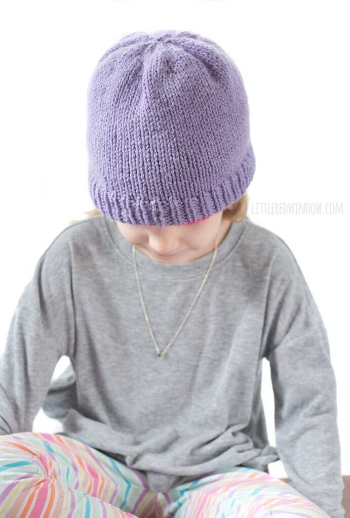 blonde girl in gray shirt looking down at her lap wearing a solid light purple knit hat with the magenta reverse color peeking out around the bottom
