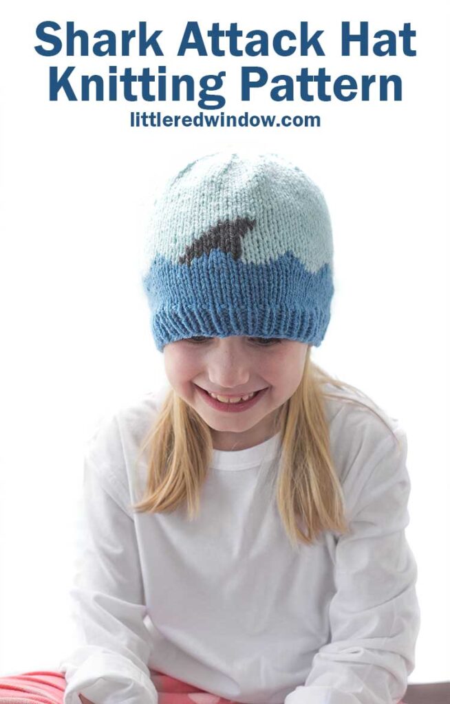 Smiling girl looking down wearing a white shirt and a knit hat with medium blue waves with a shark fin in front of a white background