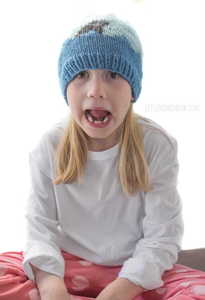 blond girl in white shirt and coral pants and the Shark Attach Hat from Little Red Window making a pretend scared face and sitting cross legged in front of a white background