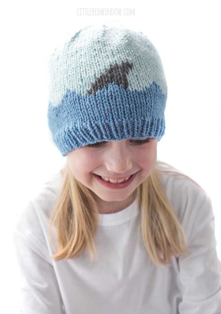Laughing girl looking down wearing a white shirt and a knit hat with medium blue waves with a shark fin in front of a white background