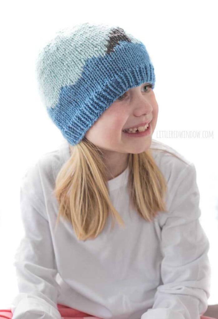Smiling girl looking off to the right wearing a white shirt and a knit hat with medium blue waves with a shark fin in front of a white background