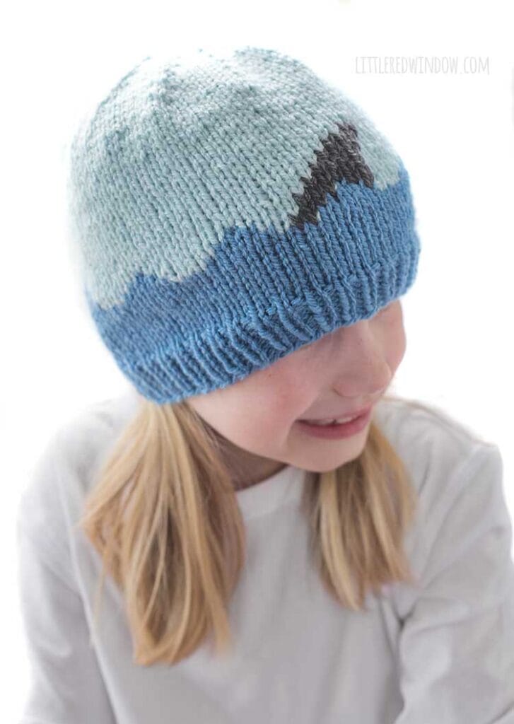 View from slightly above Smiling girl looking down and right wearing a white shirt and a knit hat with medium blue waves with a shark fin in front of a white background
