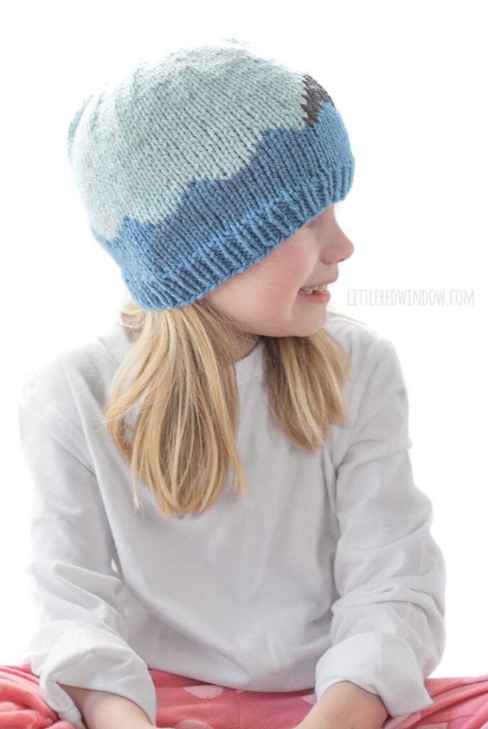 Side view of Smiling girl looking off to the right wearing a white shirt and a knit hat with medium blue waves with a shark fin in front of a white background