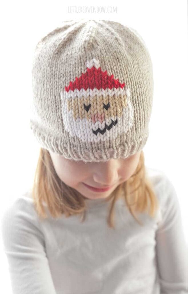 smiling girl looking down wearing a tan knit hat with a duplicate stitch Santa Face on the front of the hat with white beard smile and classic red Santa hat
