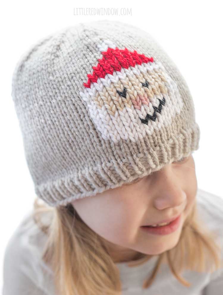 slightly above view of smiling girl looking off to the right wearing a tan knit hat with a duplicate stitch Santa Face on the front of the hat with white beard smile and classic red Santa hat