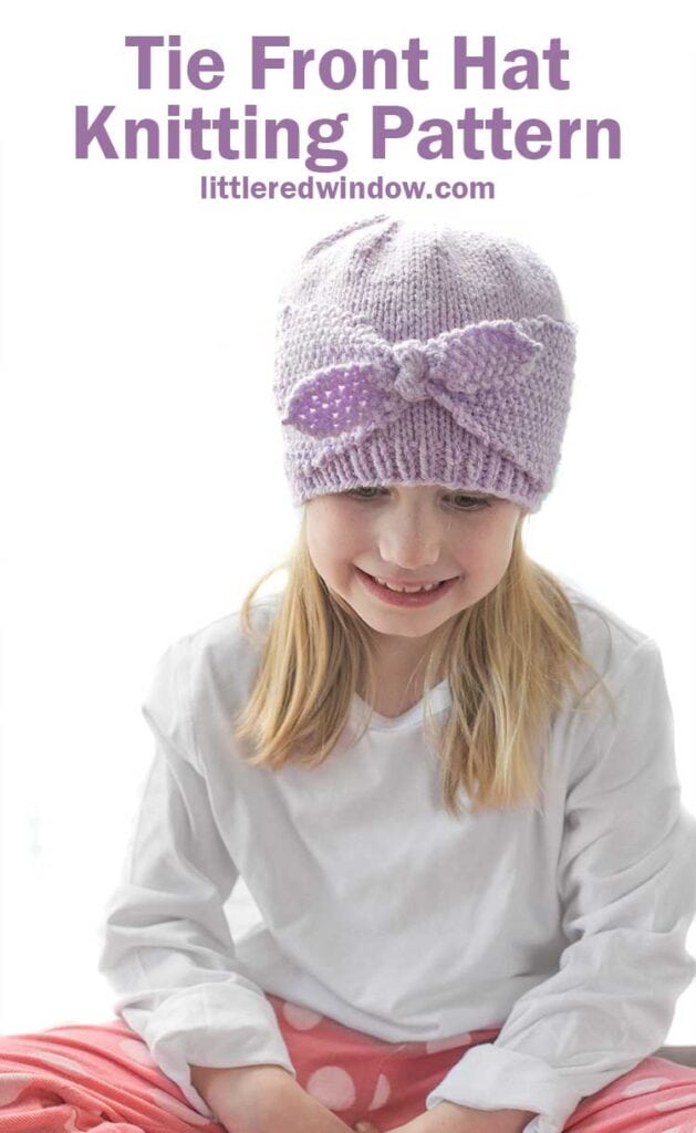 Smiling girl in white shirt and pink pants wearing a light purple knit hat with two side flaps that come together and tie in a bow in the front of the hat looking down at her lap