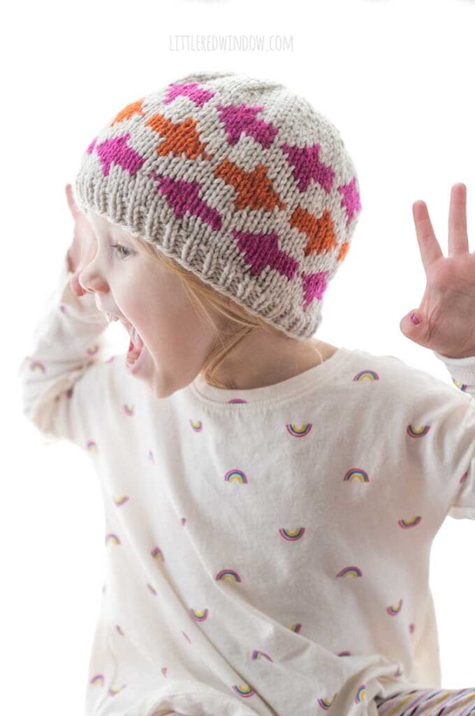 little girl roaring like a lion and wearing a tan knit hat with pink and orange arrow shapes on it