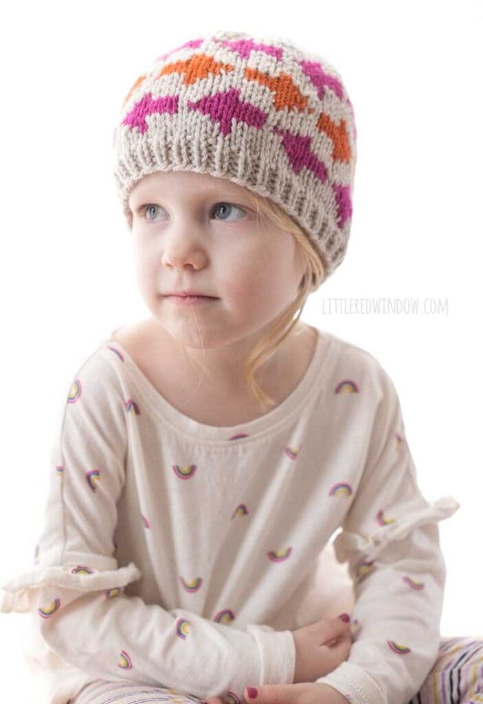 little girl wearing tan knit hat with pink and orange arrow shapes on it looking up to the left