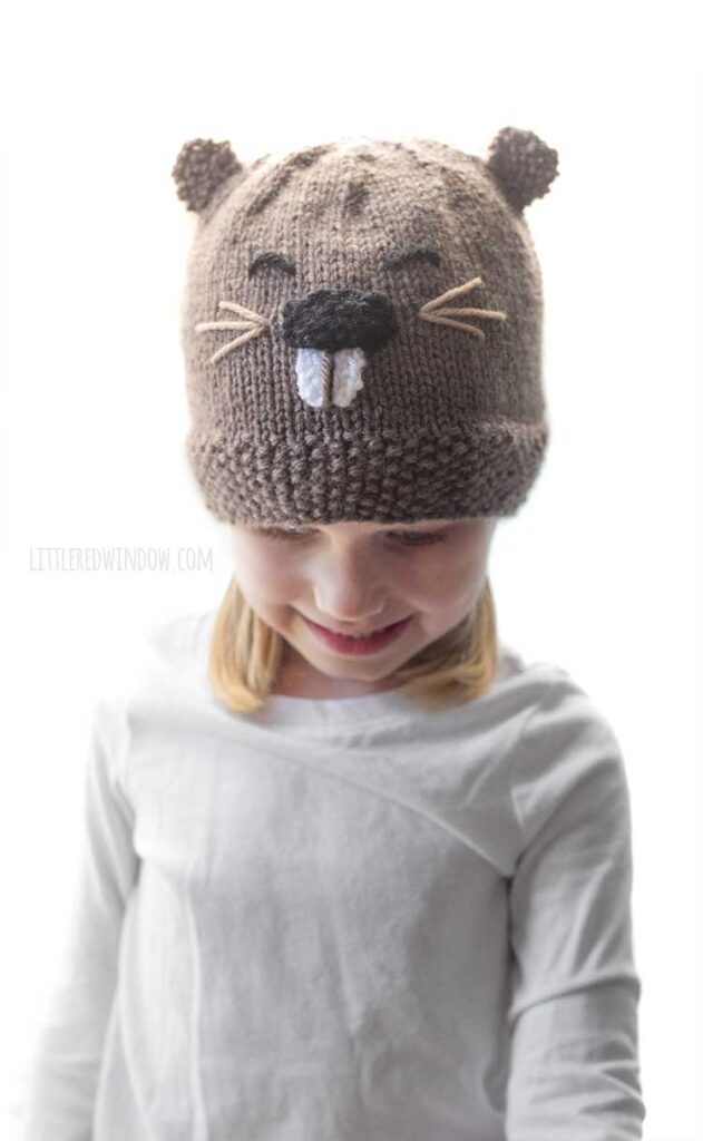 little girl wearing a brown knit hat with a beaver face and ears on the front looking down and smiling