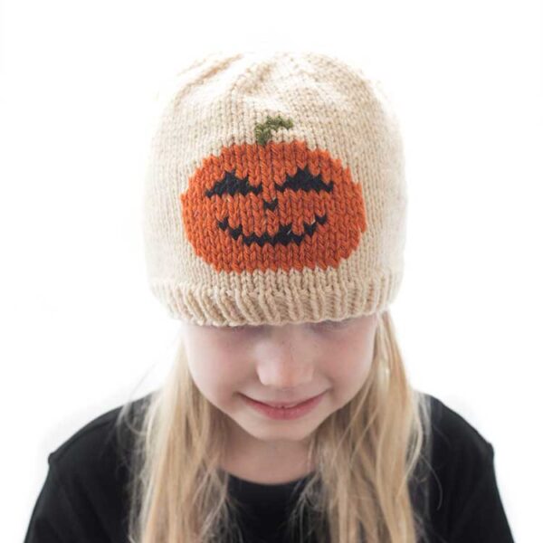 Blond girl wearing a black shirt and a tan knit hat with a smiling jack o lantern in duplicate stitch on the front center while she is smiling and looking down in front of a white background