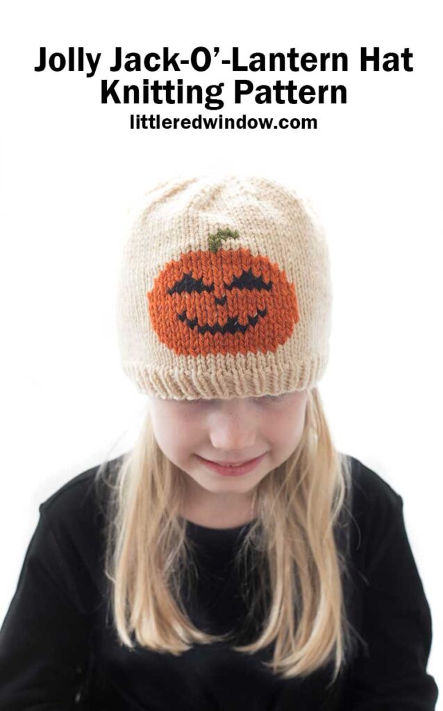 Blond girl wearing a black shirt and a tan knit hat with a smiling jack o lantern in duplicate stitch on the front center while she is smiling and looking down in front of a white background