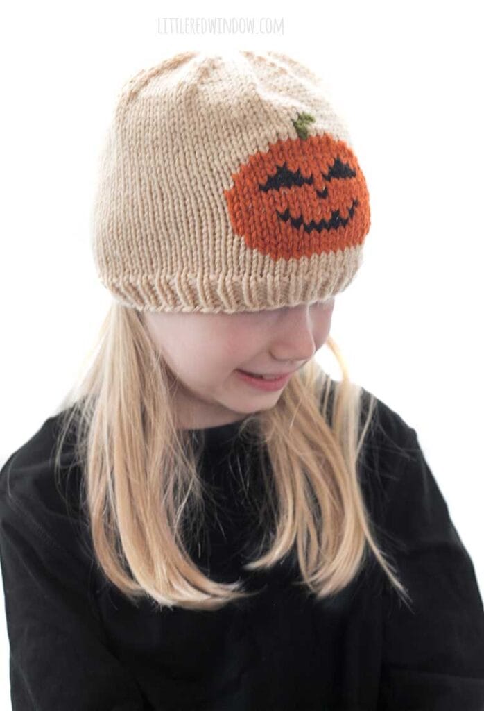 Blond girl wearing a black shirt and a tan knit hat with a smiling jack o lantern in duplicate stitch on the front center while she is smiling and looking down to the right in front of a white background