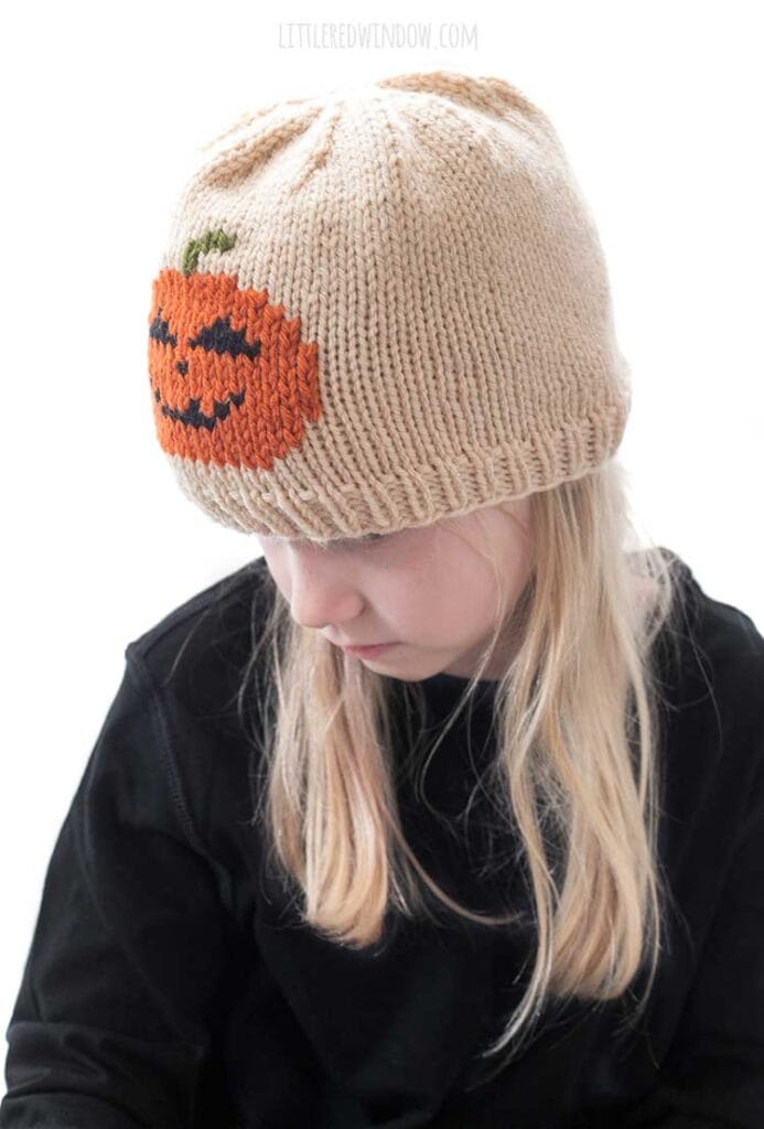 Blond girl wearing a black shirt and a tan knit hat with a smiling jack o lantern in duplicate stitch on the front center while she is looking down to the left in front of a white background
