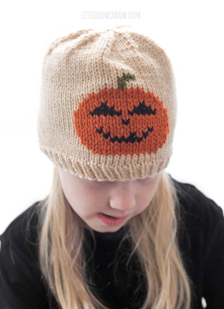 Blond girl wearing a black shirt and a tan knit hat with a smiling jack o lantern in duplicate stitch on the front center while she is looking down in front of a white background