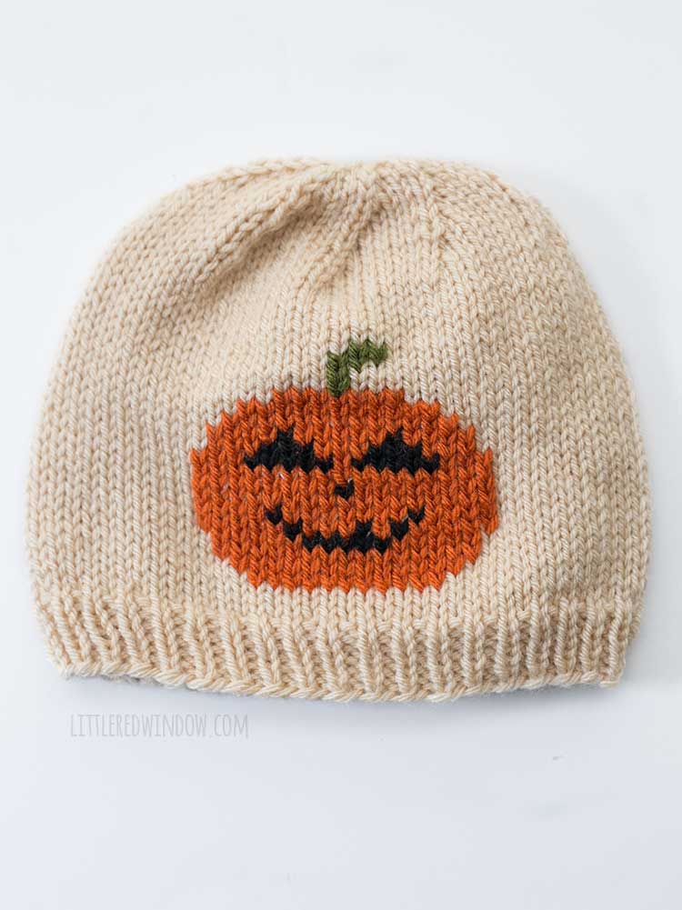 Flat lay of a tan knit hat with an orange jack o lantern in duplicate stitch on the front center in front of a white background