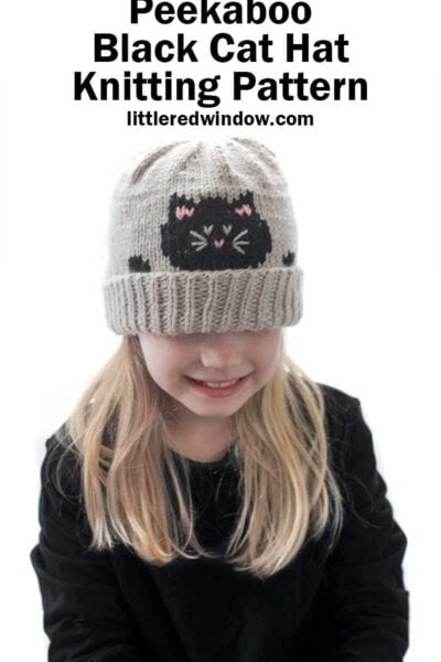 Blonde girl in a black shirt wearing a tan knit hat with a folded brim and that has a black cat face and paws peeking over the edge while she is looking down and smiling