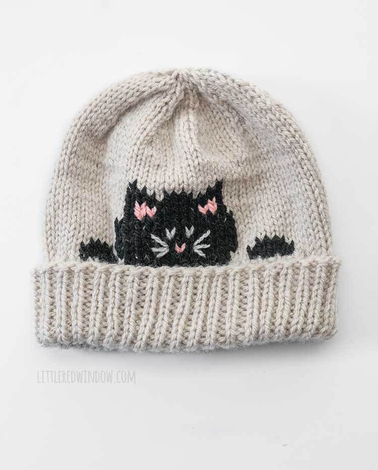 Flat lay of tan knit hat with folded ribbed brim on a white background The hat has a black cat face duplicate stitch pattern on the front peeking over the brim