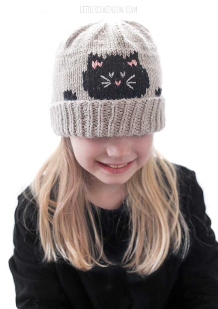 Blonde girl in a black shirt wearing a tan knit hat with a folded brim and that has a black cat face and paws peeking over the edge while she is looking down and smiling