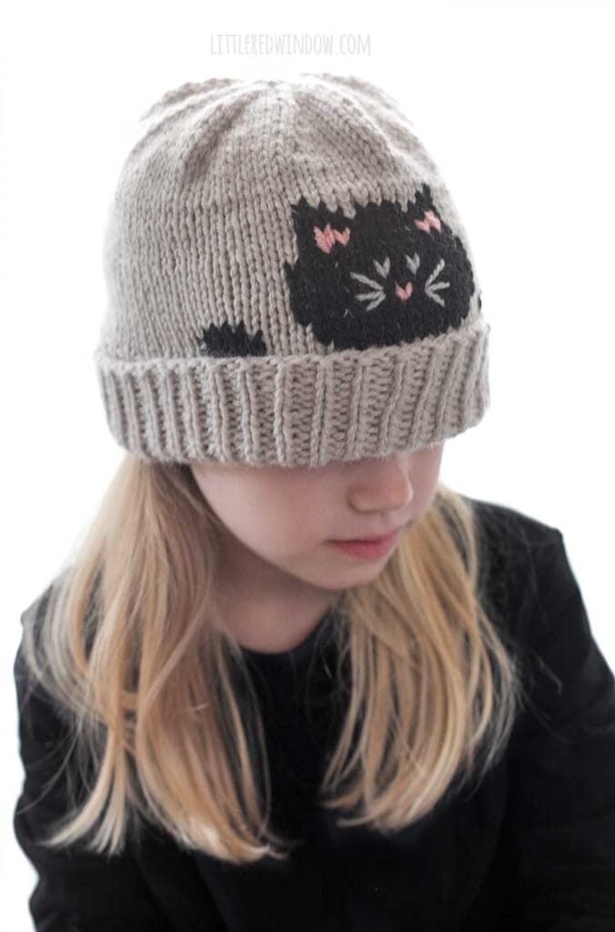 Blonde girl in a black shirt wearing a tan knit hat with a folded brim and that has a black cat face and paws peeking over the edge while she is looking down to the right and smiling