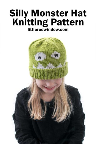 Blond girl in black shirt wearing a lime green knit hat with a monster face on the front and looking down at her lap