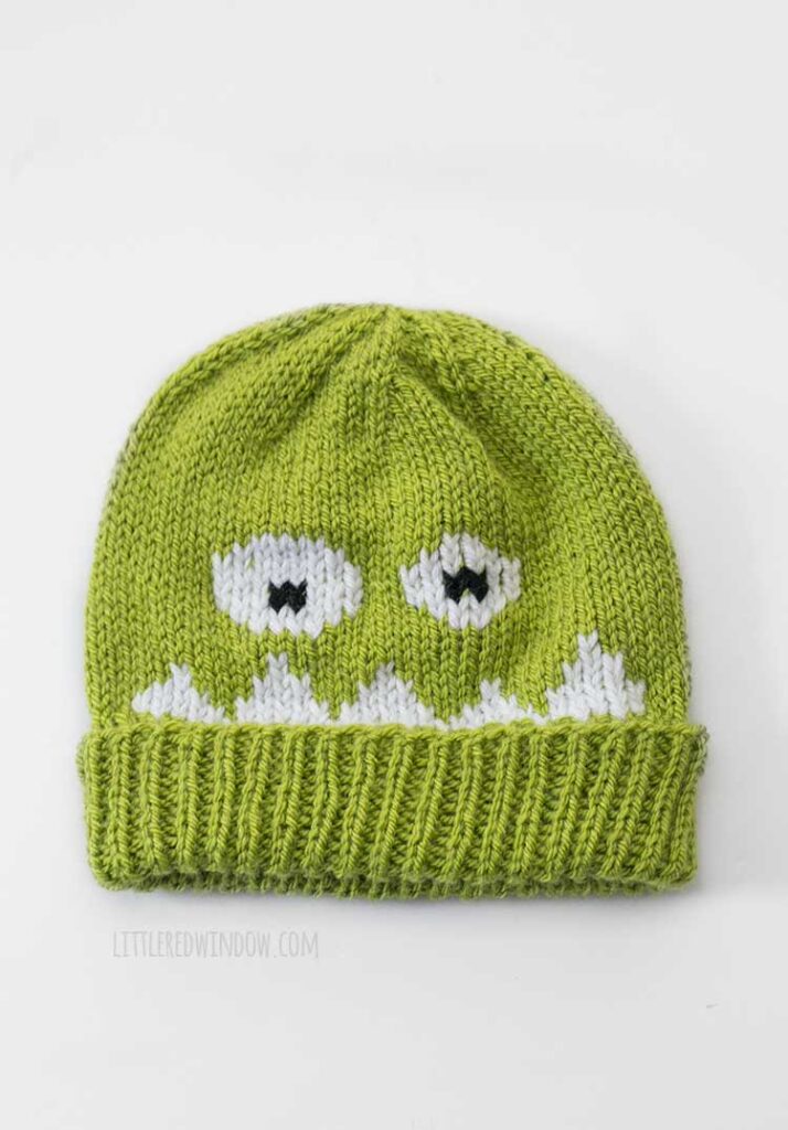 Flat lay image of a lime green knit monster hat with a folded brim on a white background the monster has white spiky teeth and two googly looking eyes on the front of the hat