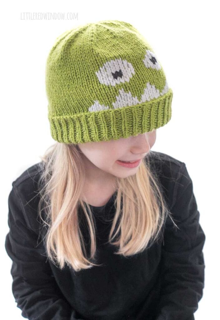 Blond girl in black shirt wearing a lime green knit hat with a monster face on the front and looking to the right and smiling