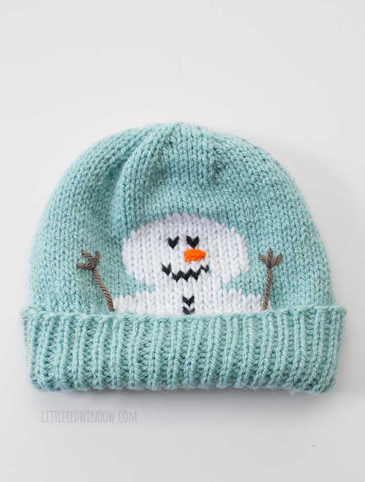 Flat lay image of a light blue knit hat with a snowman face peeking out over the folded brim in front of a white background